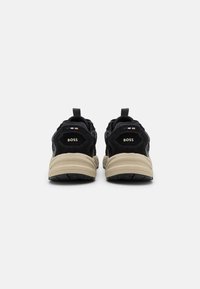 Baskets noires en suede et mesh avec une semelle beige, présentant un détail de logo à l'arrière et des languettes pour un enfilage facile.