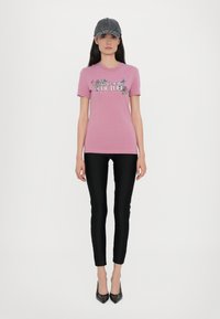 Rosa bomulls t-skjorte med "Versace Jeans Couture" grafikk, paret med svarte tettsittende leggings og svarte spisse hæler. Grå caps fullfører antrekket.