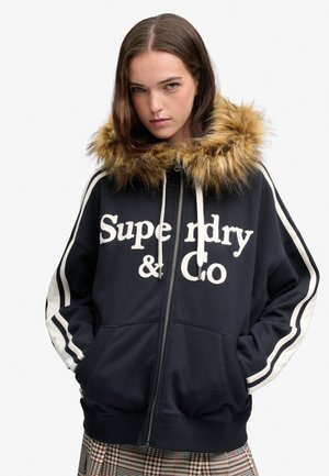 Ung kvinde iført en sort lynlåsjakke med hvide striber, pelskantet hætte og fed "Superdry & Co" tekst foran.
