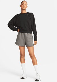 Sweatshirt noir avec des découpes, associé à un short gris. Le mannequin porte des chaussettes blanches et des baskets, affichant un look sportif et décontracté.