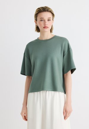 Junge Frau mit kurzen blonden Haaren, die ein lockeres grünes Kurzarmoberteil und einen weißen Rock trägt, steht vor einem einfarbigen Hintergrund.