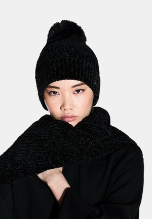Beanie nera in maglia con un pom-pom sulla parte superiore, abbinata a una sciarpa nera in maglia spessa e strutturata. Entrambi hanno un aspetto morbido e accogliente.