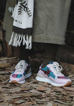 Personne portant des baskets à motifs colorés avec des lacets blancs, des chaussettes vert foncé et un pantalon beige large debout sur des feuilles sèches.