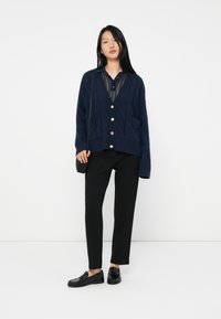 Mujer vistiendo un cárdigan azul marino abotonado sobre una blusa azul marino transparente, pantalones negros, mocasines negros y llevando un pequeño bolso negro de hombro, de pie.