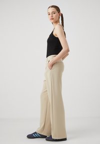 Beige, széles szárú nadrág sima anyagból, oldalsó zsebekkel és szabott derékkal, fekete ujjatlan felsővel és kék részletekkel ellátott cipővel párosítva.