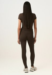 Barna bordás, rövid ujjú felső, amelyet hozzáillő, magas derekú leggings-szel párosítanak. A modell háttal áll, világos sportcipőt visel, amelynek texturált felülete van.
