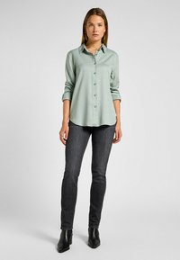 Chemise verte claire à manches longues en tissu lisse, avec un col et des manches retroussées, associée à un jean skinny foncé et des bottines noires.