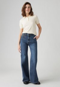 Cream short-sleeve polo shirt paired with high-waisted, wide-leg blue jeans made of denim. Model stands confidently, hand on hip.