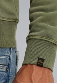 Green sweatshirt with ribbed cuffs, featuring stitching details. Black tag with "PME LEGEND" in gold attached to the cuff. Denim background visible.