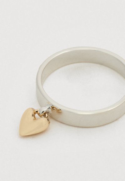 Ασημένιο δαχτυλίδι με λείο, στρογγυλό δακτύλιο και χρυσή καρδιά-γούρι. Η καρδιά έχει γυαλιστερή επιφάνεια και είναι προσαρτημένη με ένα μικρό κρίκο.