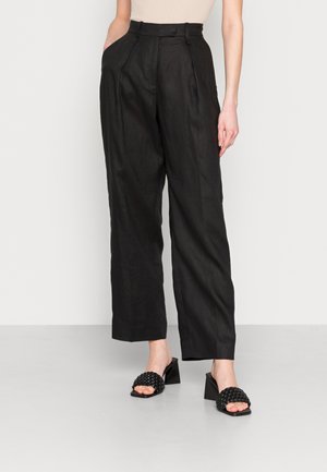 Black linen trousers with a high waist, pleated front, and side pockets, paired with woven black slip-on shoes on a concrete floor.