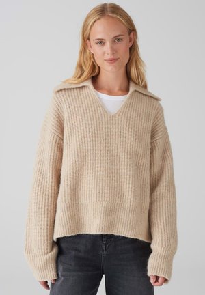 Beige knitted sweater with a wide V-neck, ribbed texture, and a cropped fit. Features long sleeves and a soft, chunky material.