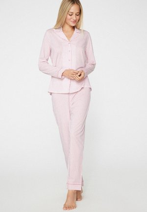 Donna con pigiama a maniche lunghe a righe rosa e bianco, in piedi scalza con le mani delicatamente intrecciate, guarda verso il basso e sorride dolcemente.