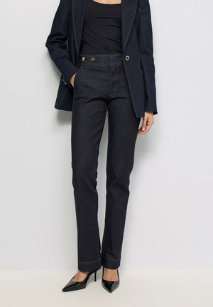 Femme portant un jean sombre ajusté, un top noir, un blazer sombre et des talons aiguilles noirs pointus, debout devant un fond clair uni.