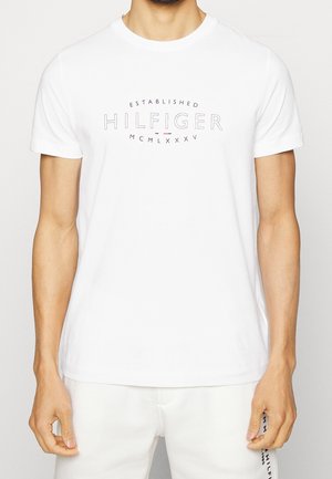 Mand iført en hvid t-shirt med korte ærmer med teksten "Established Hilfiger MCMLXXXV" trykt i sort på brystet.