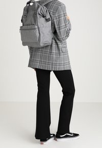 Personne portant un pantalon noir, des baskets noires et blanches, un blazer à carreaux gris et un grand sac à dos gris sur le dos.