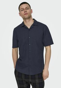 Camisa de manga corta, de color azul oscuro, con botones y hecha de una tela texturizada, que presenta un cuello de tipo camp y un corte recto con sutiles detalles de costura.