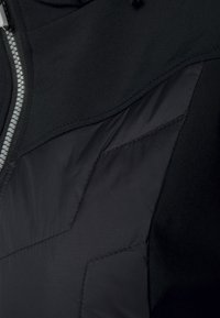 Czarna kurtka wykonana z mieszanki gładkich i teksturowanych tkanin, z zapięciem na zamek błyskawiczny i wyraźnymi przeszyciami tworzącymi wzory na materiale.