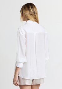 White shirt with a relaxed fit, long sleeves, and a back seam detail. Made of light, textured fabric. Simple collar design.