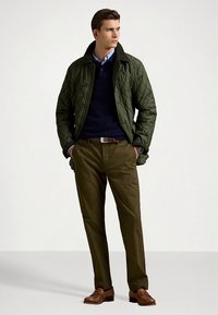 Grön quiltad jacka med krage, marinblå tröja under, brunt bälte, olivgröna byxor och bruna skor. Modell poserar mot en neutral bakgrund.