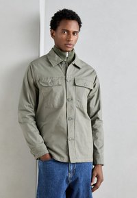 Jachetă button-up de culoare verde măslinie deschisă, cu două buzunare pe piept, mâneci lungi și croială dreaptă, purtată cu blugi albaștri. Finisaj din țesătură texturată.