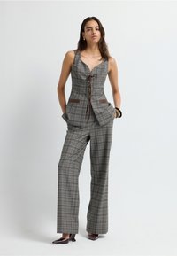 Mono de cuadros gris a medida con un cuerpo ajustado, escote abierto, frente con cordones y detalles de cuero marrón. Pantalones de pierna ancha y bolsillos laterales.
