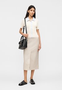 Γυναίκα ντυμένη με μπεζ κοντομάνικo πουκάμισo με κουμπιά, μπεζ μίντι φούστα, μαύρα μοκασίνια και κρατώντας μια μαύρη τσάντα στον ώμο της.