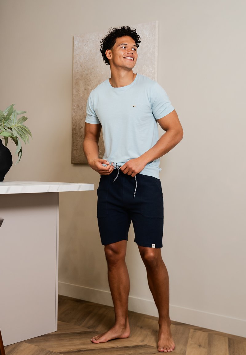 Světle modré tričko s krátkým rukávem a malým emblémem, v kombinaci s tmavě modrými šortkami s pružným pasem a bočními kapsami.