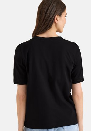 Frau, die mit dem Rücken zur Kamera steht, trägt ein schlichtes schwarzes Kurzarmshirt und hellblaue Jeans vor weißem Hintergrund.