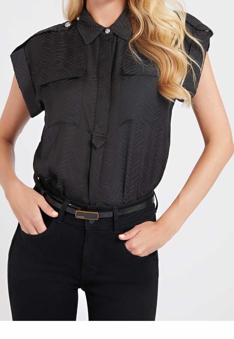 Schwarzes Hemd mit kurzen umgeschlagenen Ärmeln, das ein strukturiertes Fischgrätenmuster aufweist und zwei Brusttaschen hat. Getragen mit schwarzen Hosen und Gürtel.