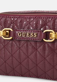 Bordeauxfarbenes, gestepptes Täschchen aus glänzendem Material, mit goldenen Hardware-Akzenten und einer gesteppten Verzierung, sowie einem sichtbaren Logo auf der Vorderseite.