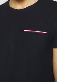 Schwarzes T-Shirt mit rundem Ausschnitt, kurzen Ärmeln und einer Brusttasche, die mit einem rot-weißen und blauen gestreiften Akzent versehen ist. Strukturiertes Material.