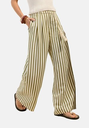 Person iført brede bukser med grønne og hvide vertikale striber, beige ærmeløs top, sandaler og træarmbånd, hånd i lommen, går.
