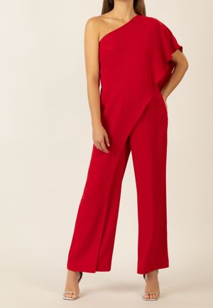 Femme portant une combinaison rouge à une épaule avec jambes larges et des talons ouverts argentés, debout avec une main sur la hanche.