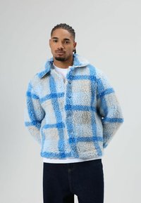 Light blue and cream plaid fleece jacket with a collar, visible buttons, and a soft, textured surface. Worn over a white shirt.