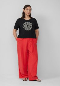 Zwarte cropped t-shirt met witte geometrische print, gecombineerd met losse rode wijde broek. Model staat met handen in de zakken tegen een neutrale achtergrond.