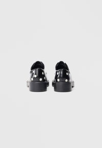 Sorte sko i laklæder med store hvide polkaprikker, rundet snude, flad sål og minimale syningsdetaljer på bagsiden.