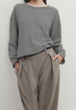 Personne portant un chemisier à manches longues rayé noir et blanc, rentré dans un pantalon beige plissé taille haute, les mains dans les poches.