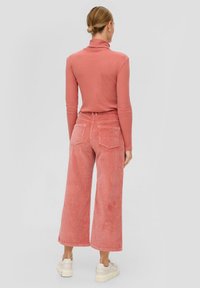 Woman with blonde hair in a bun wearing a coral turtleneck and matching wide-leg corduroy pants, standing with back to the camera.