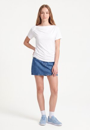 Jonge vrouw met lang haar die een wit topje met korte mouwen, een spijkerrok, witte sokken en lichtblauwe sneakers draagt, staande met één hand op haar heup.