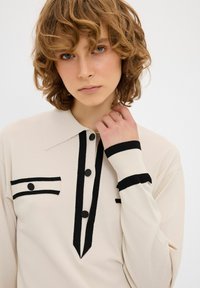 Maglione color crema con colletto a righe nere, patta con bottoni e dettagli sulle tasche. Tessuto strutturato con polsini a costine.