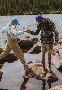 Άνδρας με μπουφάν με μοτίβο και σορτς βοηθά γυναίκα με ολόσωμη φόρμα και καπέλο να περάσει πάνω από βράχια σε μια λίμνη περιτριγυρισμένη από δάσος.