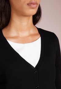 Czarny kardigan z dekoltem w serek, z teksturowaną białą bluzką pod spodem. Kardigan ma prążkowane krawędzie i czarne guziki.