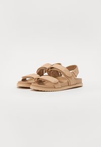 Tan sandalen met een kruisband ontwerp, een gepolsterde textuur en een platte rubberen zool met een getande rand voor grip.