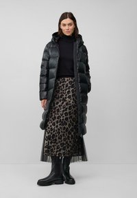 Zwarte pufferjack met een capuchon, lange snit, gedragen over een leopardprint rok en een zwarte aansluitende coltrui. Zwarte kniehoge laarzen.