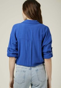Chemise légère bleue avec des manches retroussées, détail de pince dans le dos et un col, associée à un jean en denim bleu clair. Texture unie.