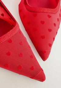 Pantofi roșii cu vârf ascuțit, dintr-un material texturat care prezintă mici modele în formă de inimă ridicate pe un fundal deschis la culoare.