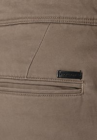 Beige Hosen mit glatter Textur, ausgestattet mit einer Gesäßtasche und einem schwarzen Etikett mit "JACK & JONES". Sichtbare Nähte in einer helleren Farbe.