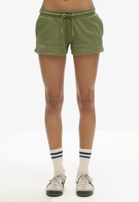 Pantalones cortos verdes de algodón con cinturilla elástica, cierre de cordón y bolsillos frontales. Detalle notable del logo en bronce en el lado izquierdo.