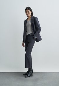 Blazer et pantalon gris sur mesure avec un haut côtelé gris clair. Bottines noires à bout pointu. Tissu lisse et coupe structurée.